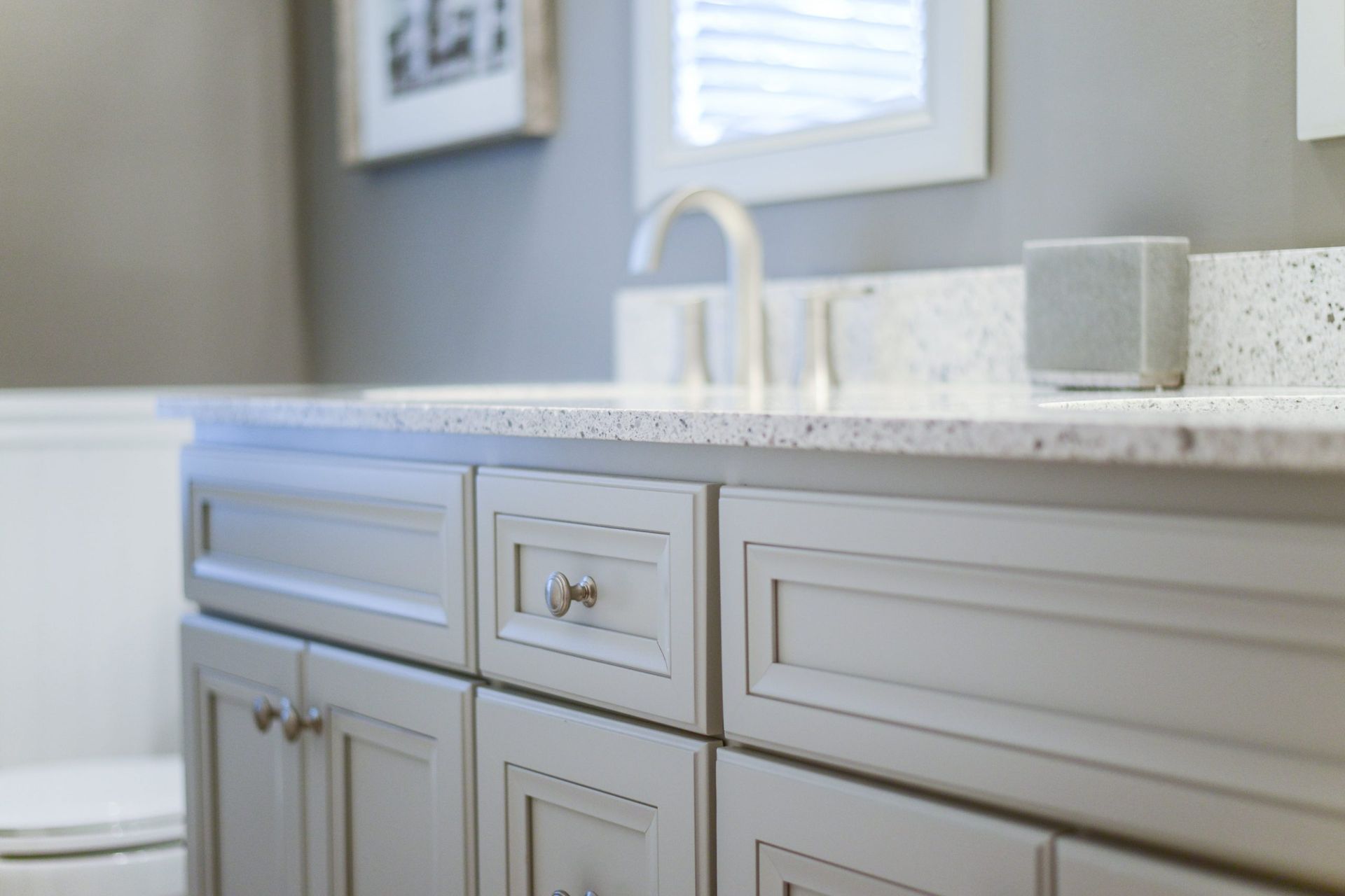 A light gray bathroom vanity with a speckled white countertop, chrome faucet, and a small speaker, against a gray wall.