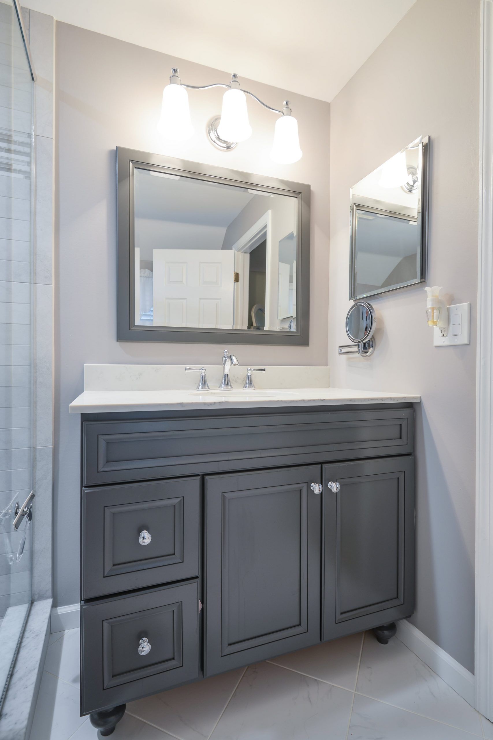 A gray bathroom vanity with a white countertop, rectangular mirror, and three-light fixture on a light gray wall.
