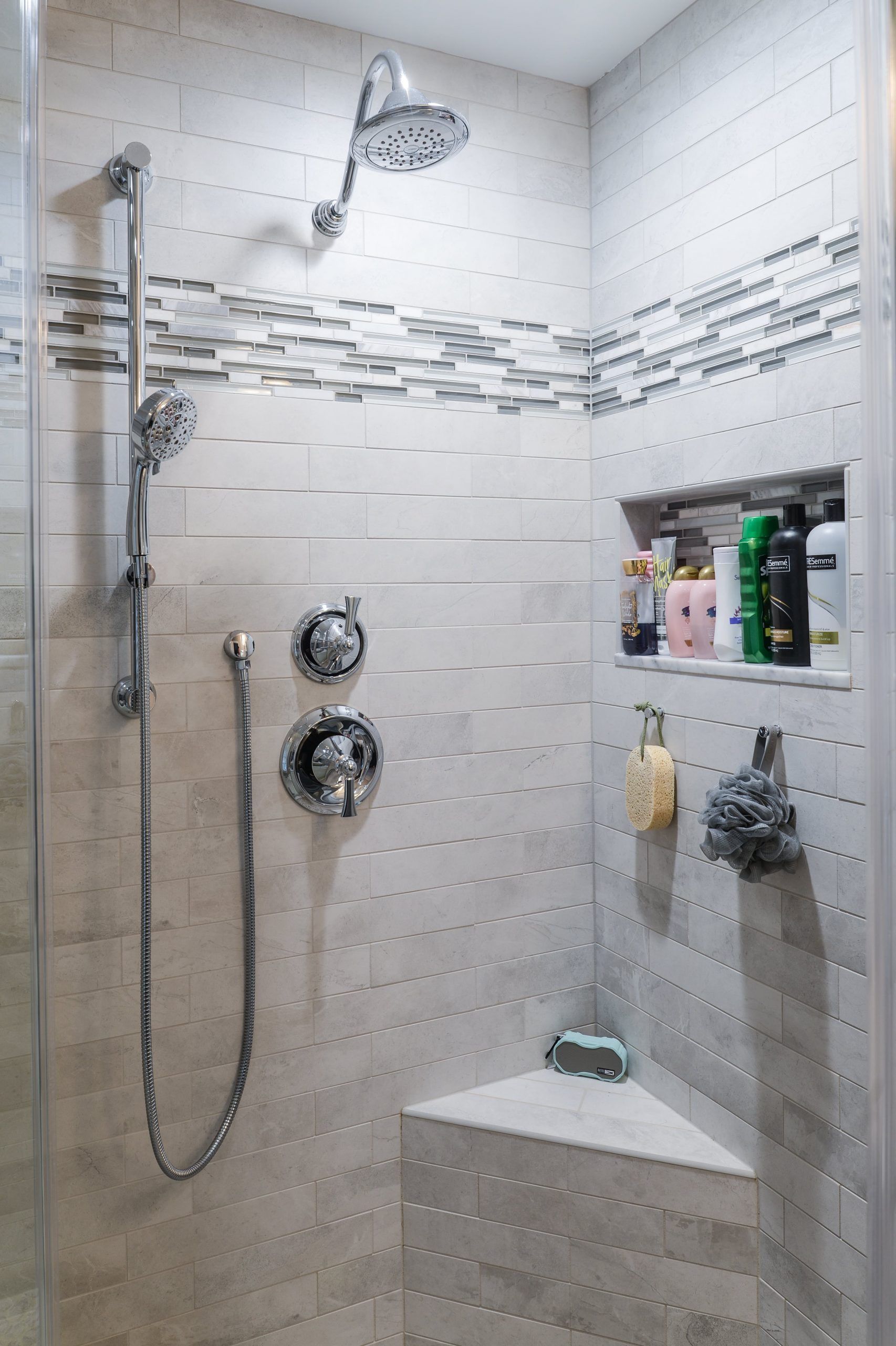 A modern tiled shower featuring a rain showerhead, handheld sprayer, built-in shelving with toiletries, and a corner seat.