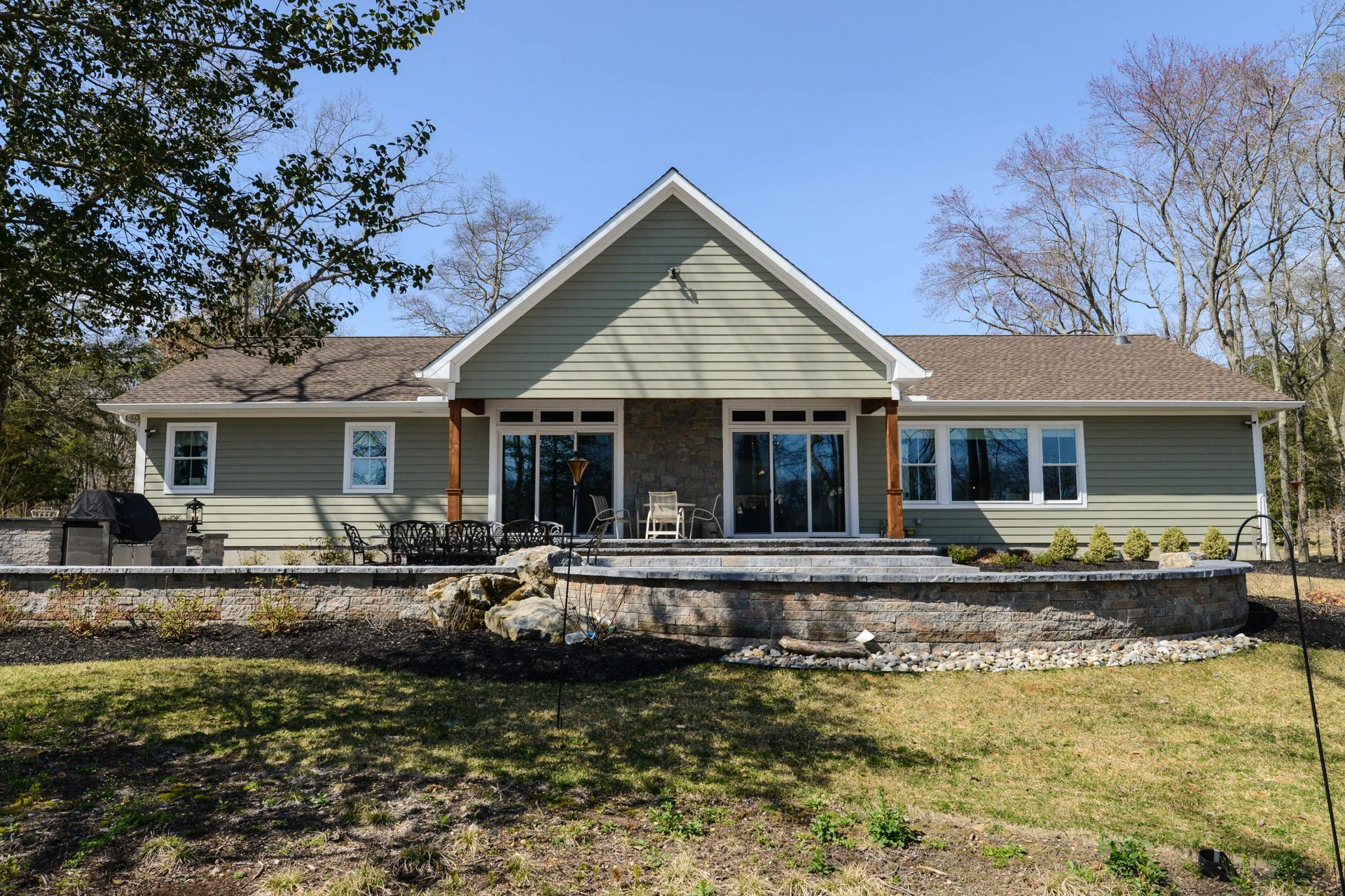 A single-story, sage green house with a stone patio, gabled roof, and glass sliding doors, surrounded by trees on a sunny day.