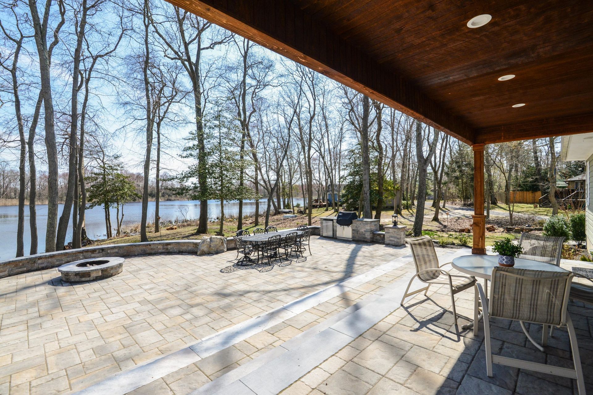 A stone patio with a fire pit and grill overlooks a lake surrounded by bare trees under a bright blue sky.