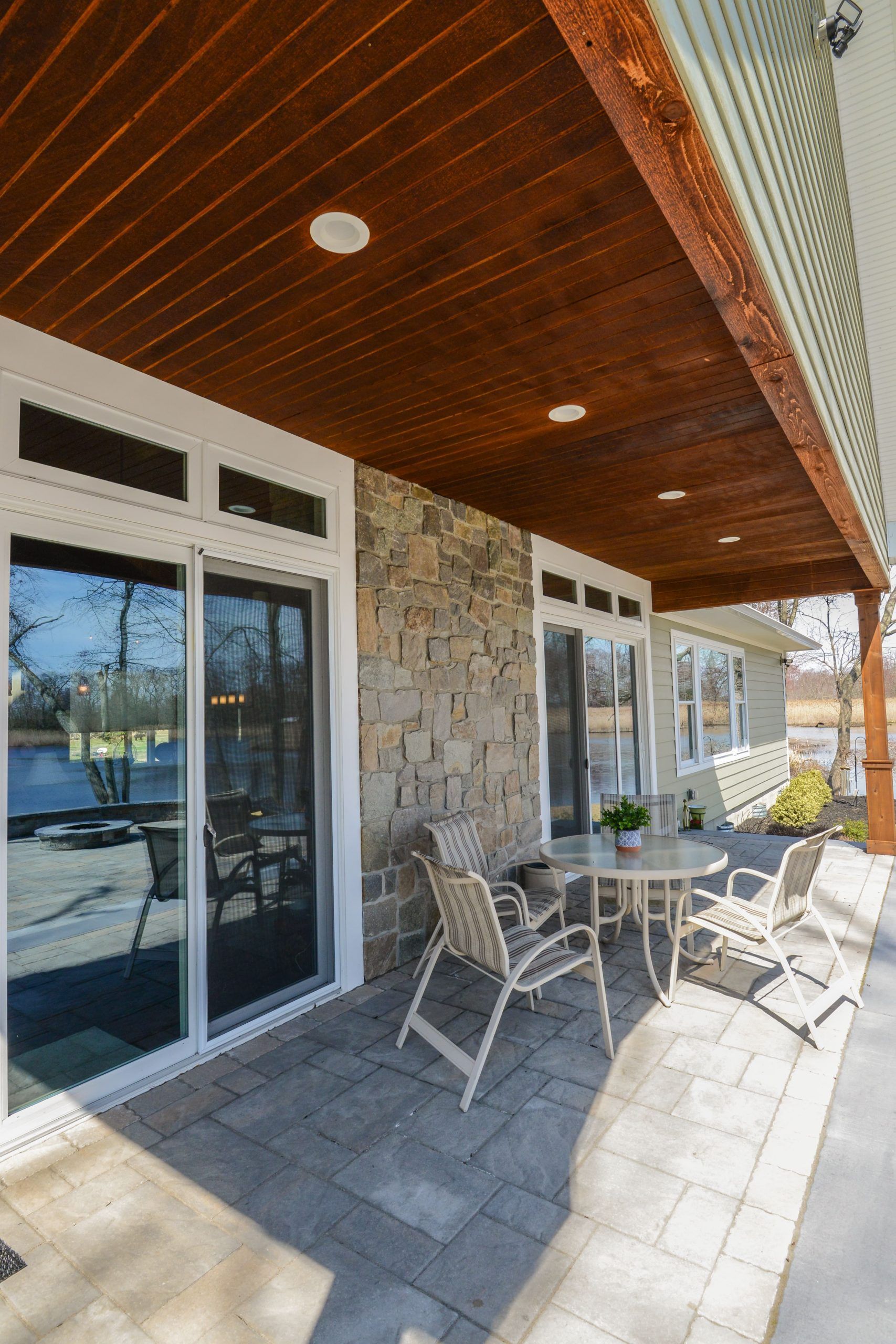 Patio with stone wall, wooden ceiling, glass sliding doors, and outdoor table and chairs overlooking a lake.