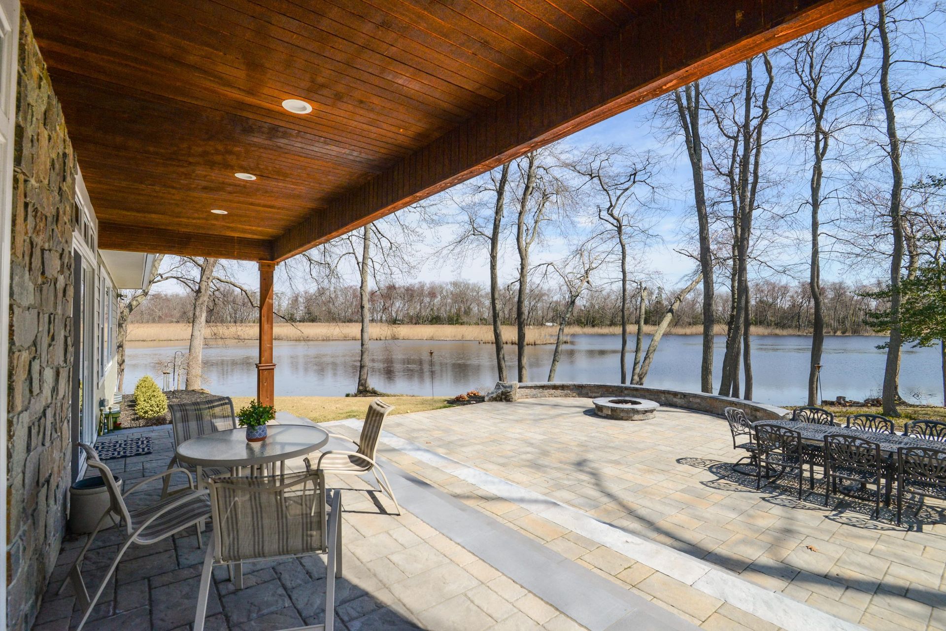 A stone patio overlooks a calm lake under a bright blue sky, shaded by a wood-paneled ceiling with outdoor seating.