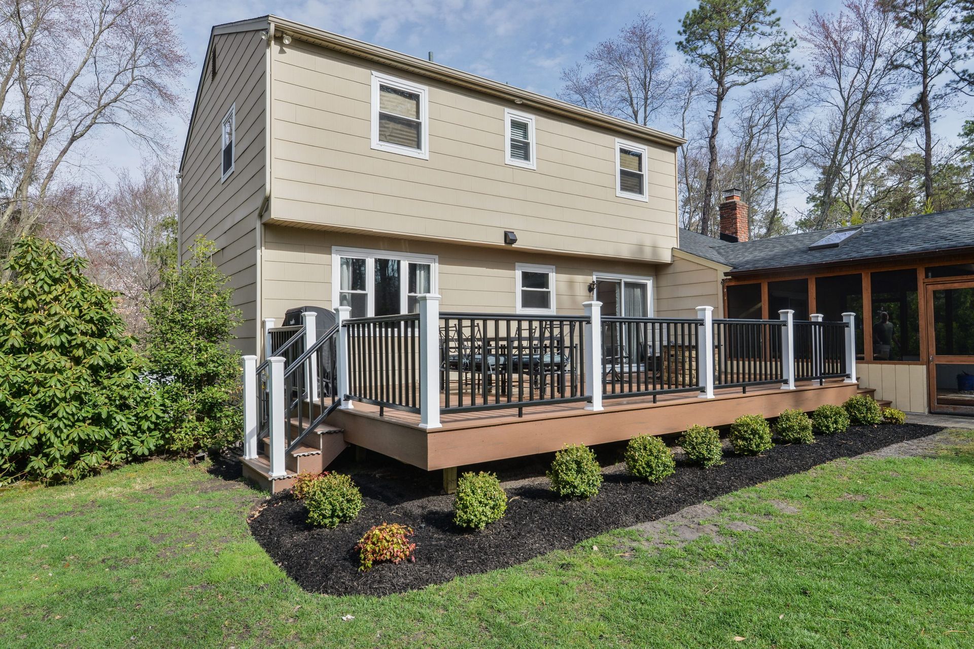 A tan two-story house with a wooden deck and black railings, situated in a backyard with landscaping and a sunroom.