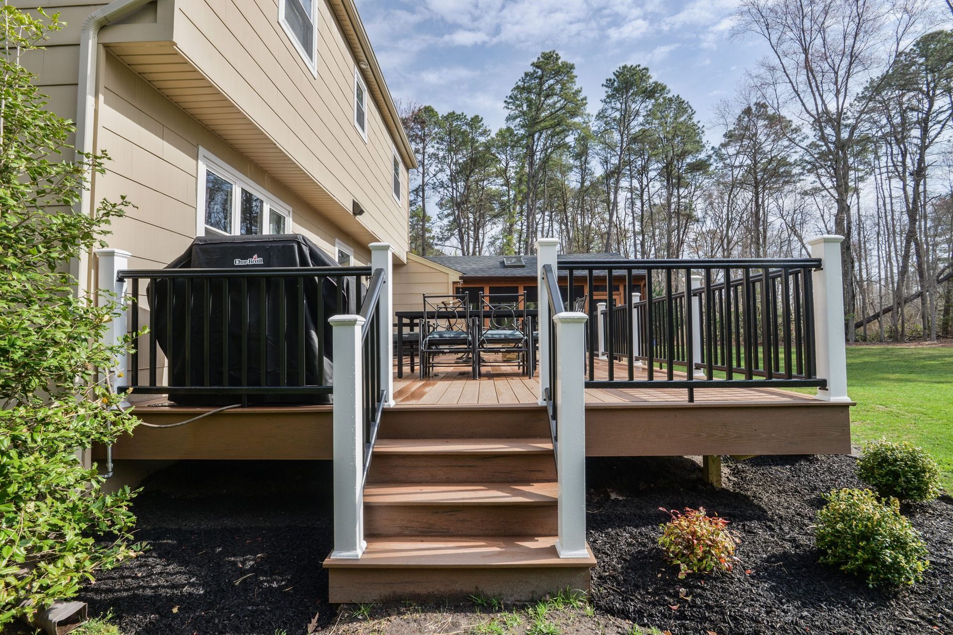 A brown wooden deck with black railings and a grill next to a tan house, overlooking a grassy yard with trees.