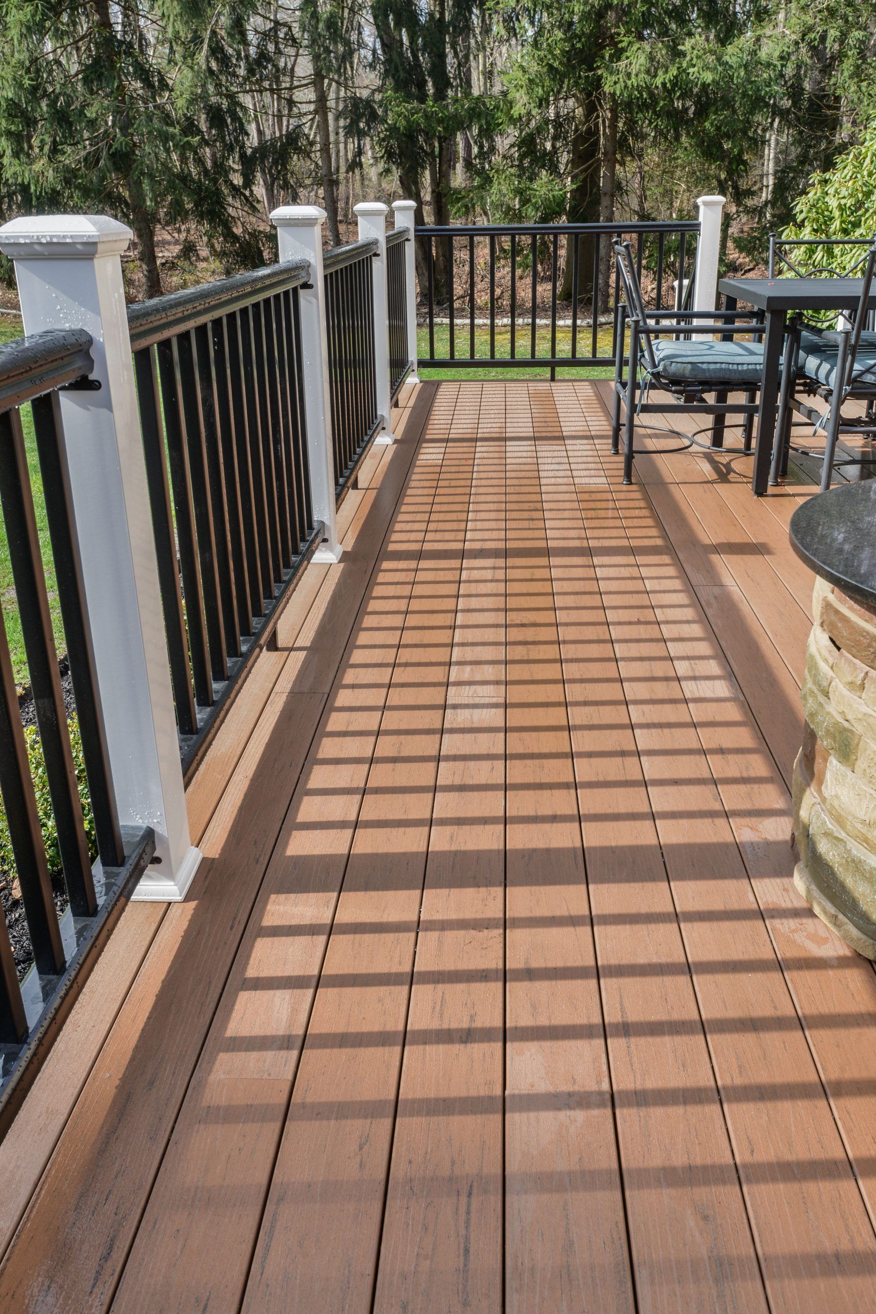 A wooden deck with black railings and white posts, featuring a matching border design and outdoor seating in the background.