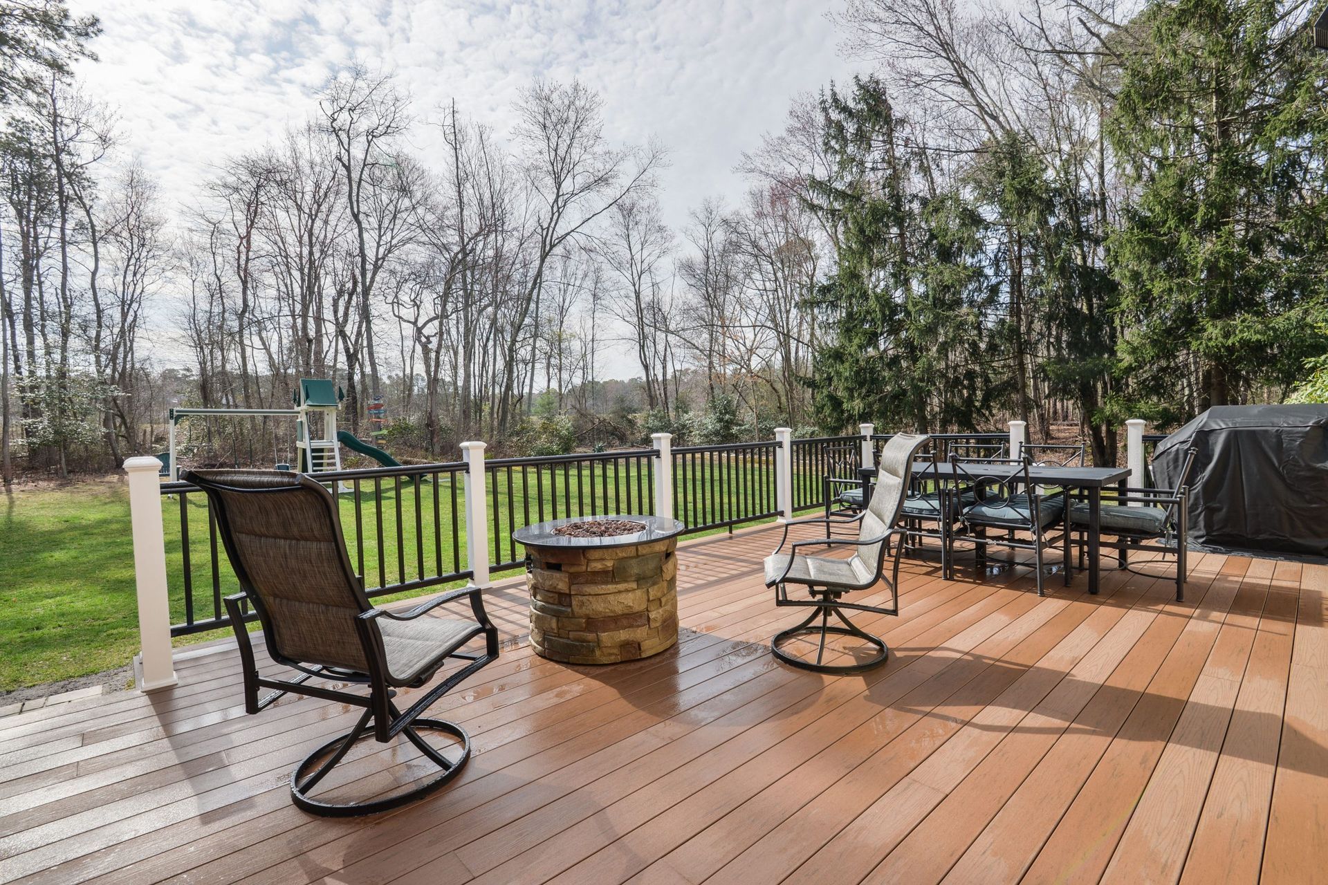 A wooden deck with a fire pit, patio chairs, a dining set, and a railing overlooking a grassy backyard and woods.