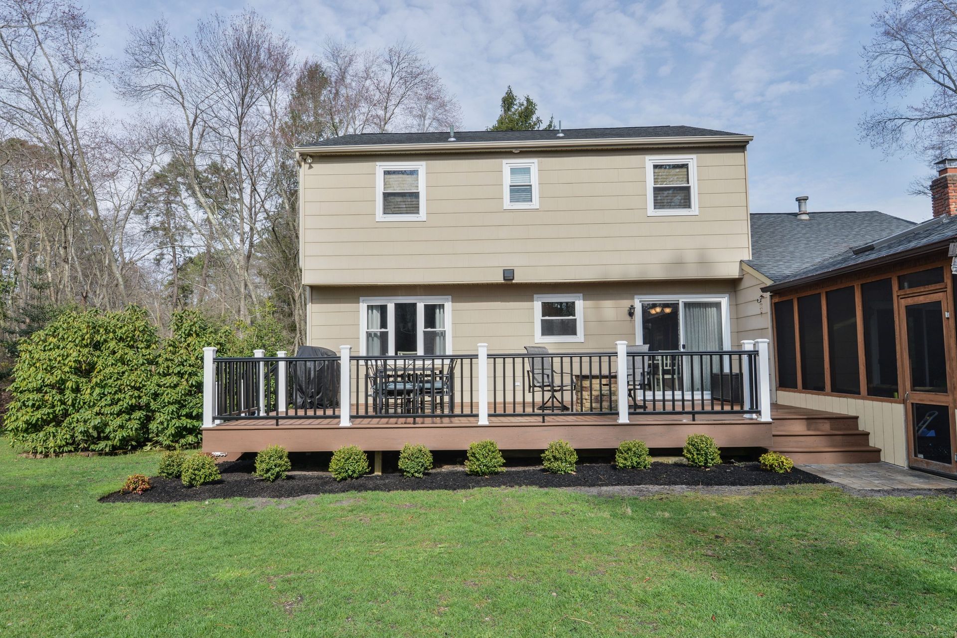 A two-story tan house with a dark wooden deck, white railings, and a screened-in porch, set against a green lawn and trees.