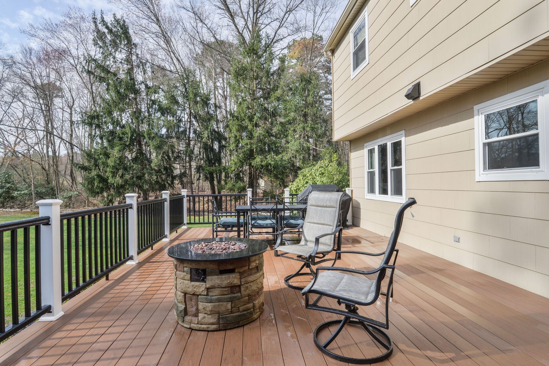 A raised deck with a stone fire pit, outdoor furniture, and a black railing overlooking a yard with trees.
