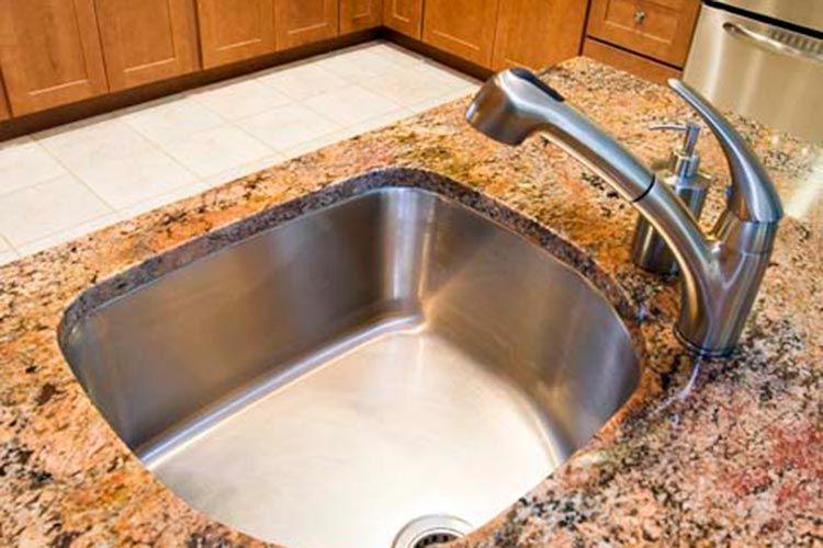 A stainless steel kitchen sink set into a speckled, orange-toned granite countertop with a brushed metal faucet.