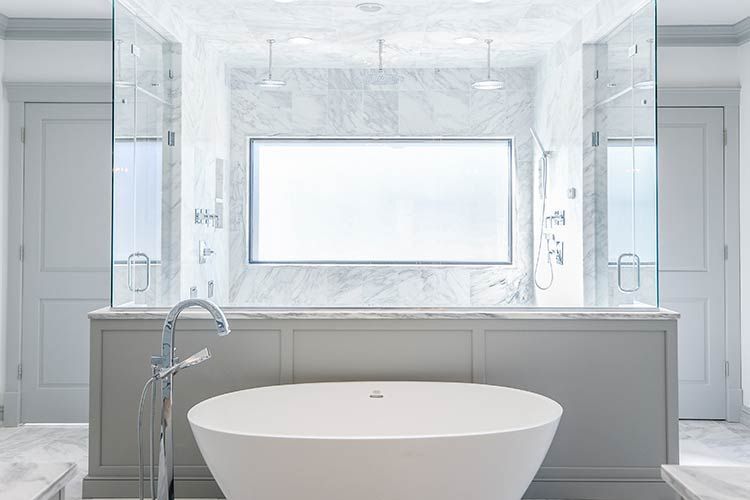 A white soaking tub sits in front of a glass-enclosed shower with marble tile and gray walls in a modern bathroom.