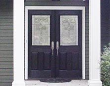 A dark double door with glass panels featuring leaded detailing, set between two white columns against gray siding.