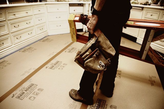 A person standing in a white kitchen holds a brown canvas tool bag while working on floor renovation.
