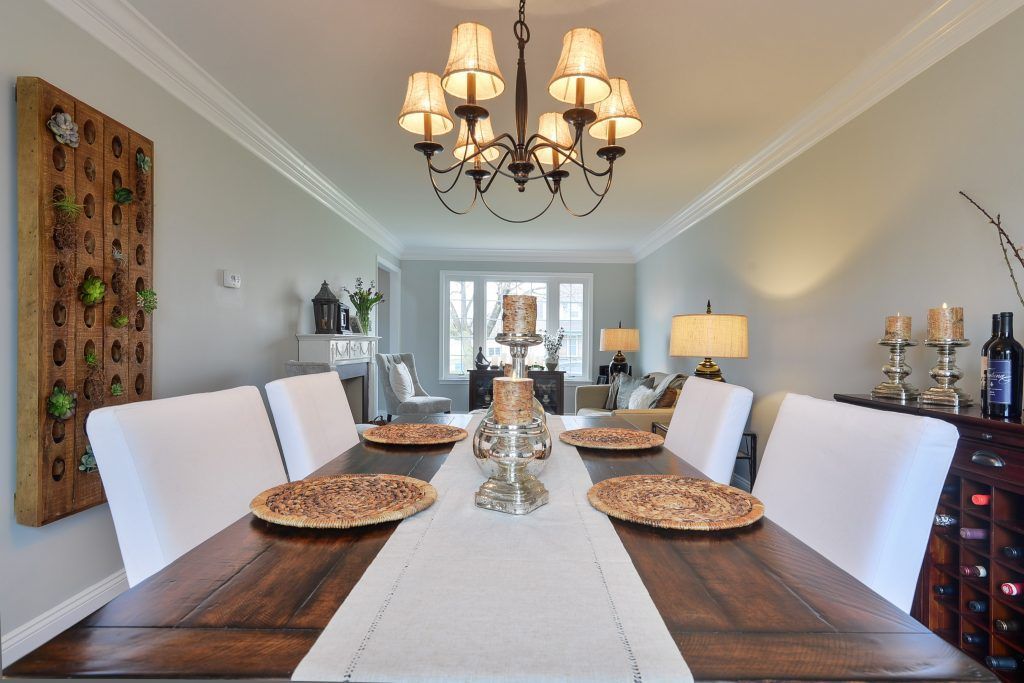 A dining room with a wooden table, white chairs, a decorative wall piece, and a chandelier, looking toward a living area.