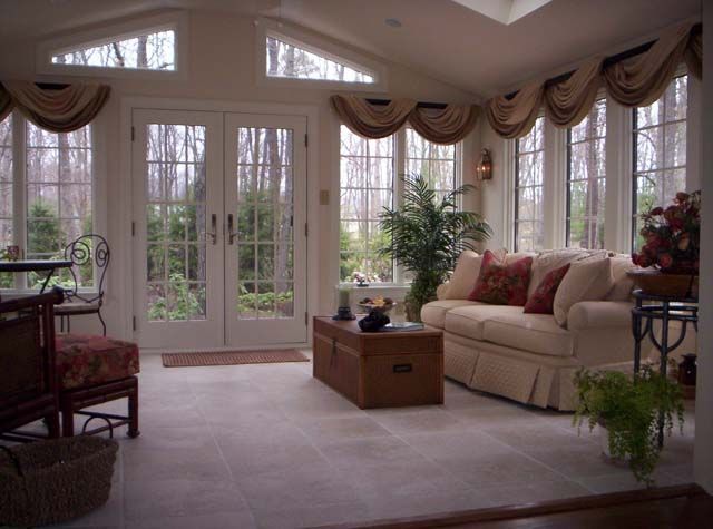 Sunroom with a beige sofa, central wooden coffee table, large windows with valances, and glass double doors.