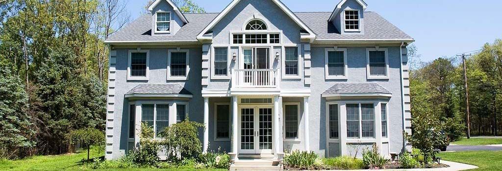 A two-story, light blue residential home with a central entrance, white trim, and a dormer-style roof in a wooded setting.