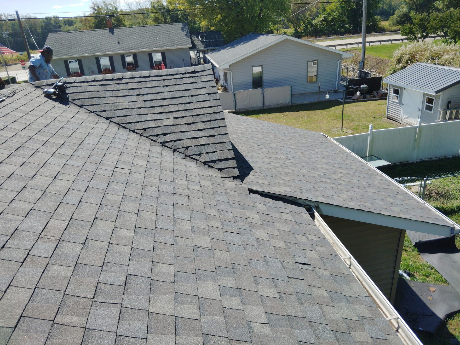 Roofer on a residential roof with gray shingles; other houses and a yard are in the background.
