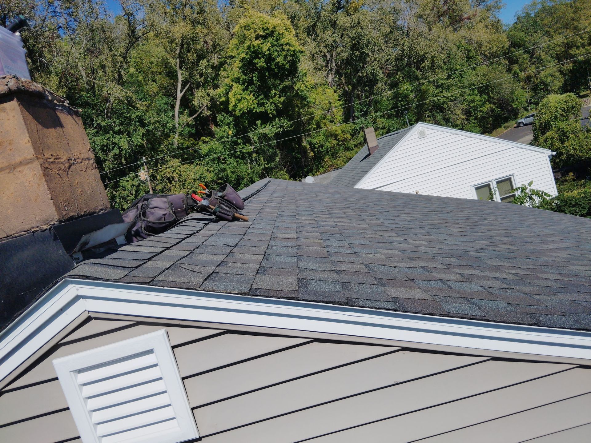 Workers repairing a shingled roof. House exterior with chimney, trees, and a white house in background.
