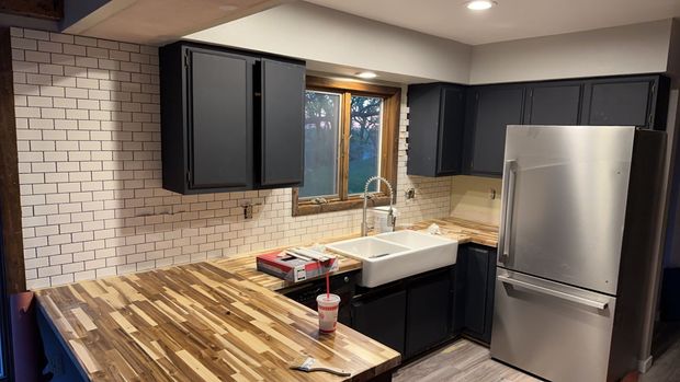 Kitchen with black cabinets, butcher block countertops, white backsplash, and stainless steel refrigerator.