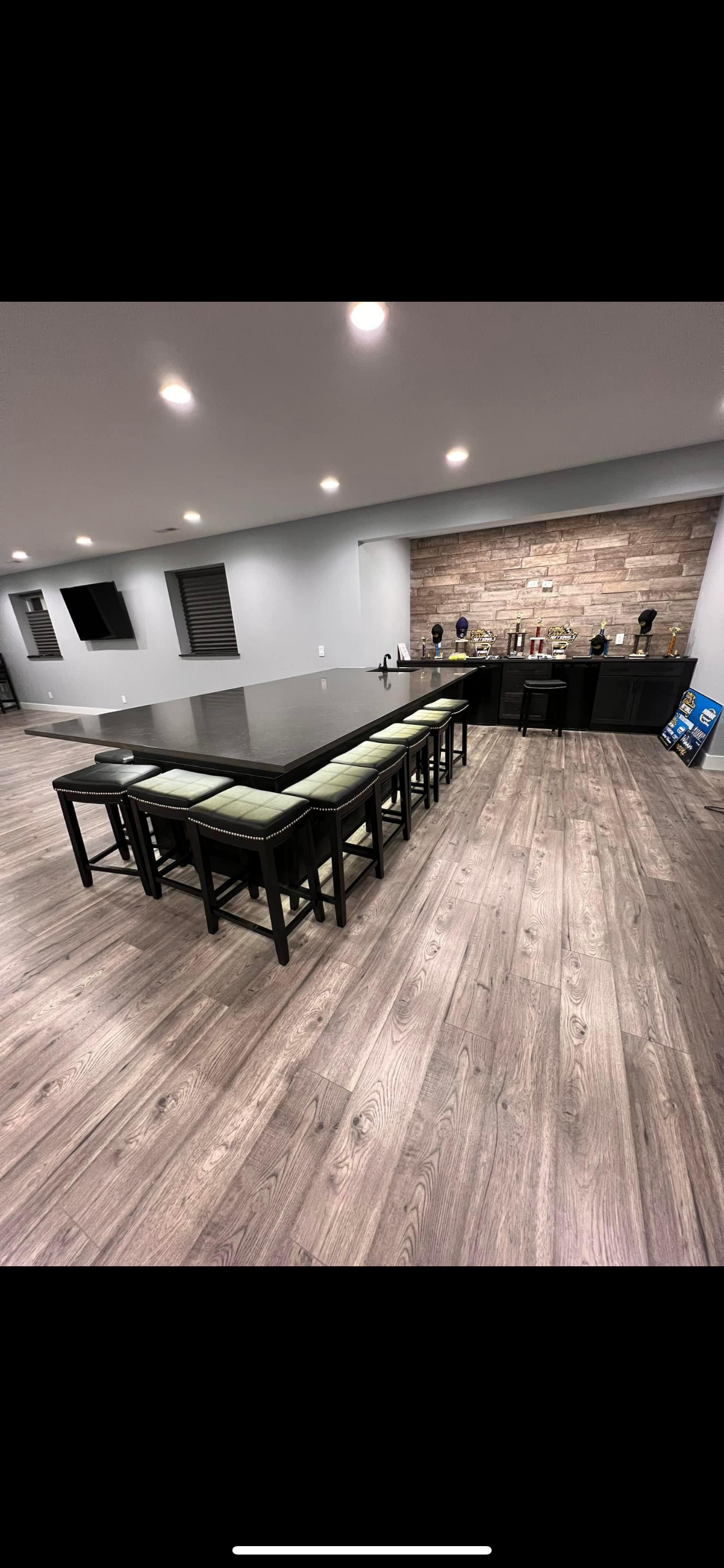 A large, dark table surrounded by stools in a room with wood-look flooring and a brick wall.