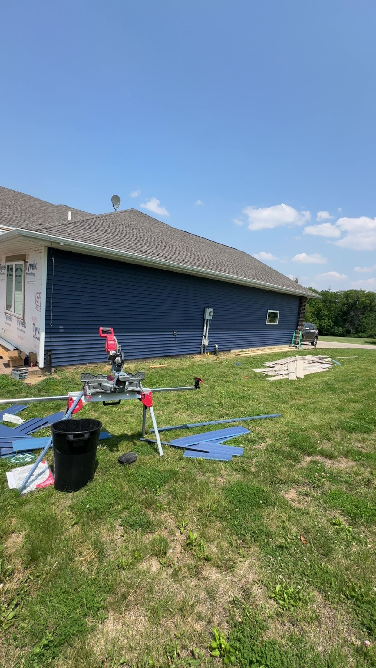 Side of a house with dark blue wrap, construction tools on grass, sunny day.