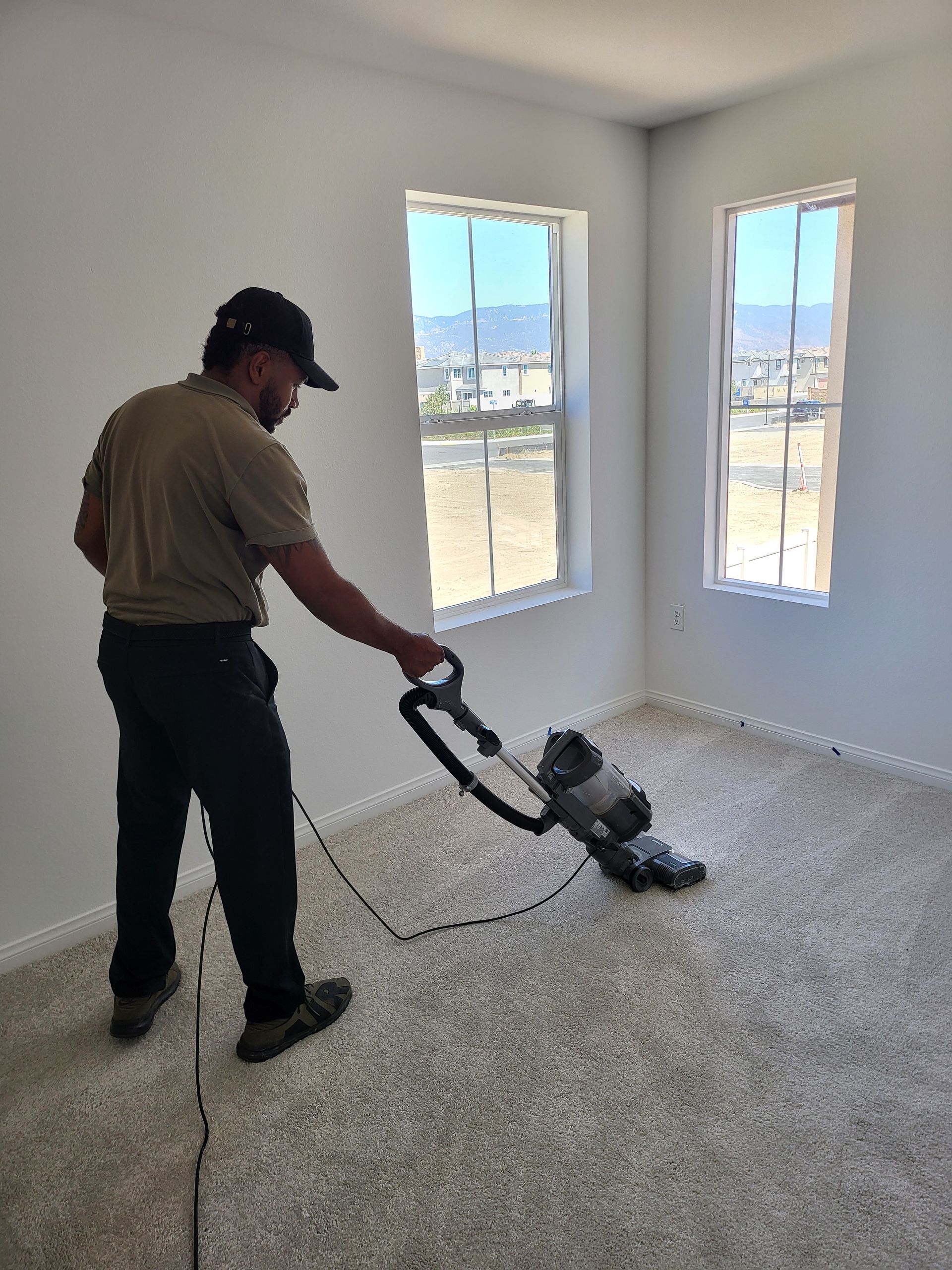 Two people cleaning an office. One vacuums the carpet, the other dusts. Large windows and modern furniture.