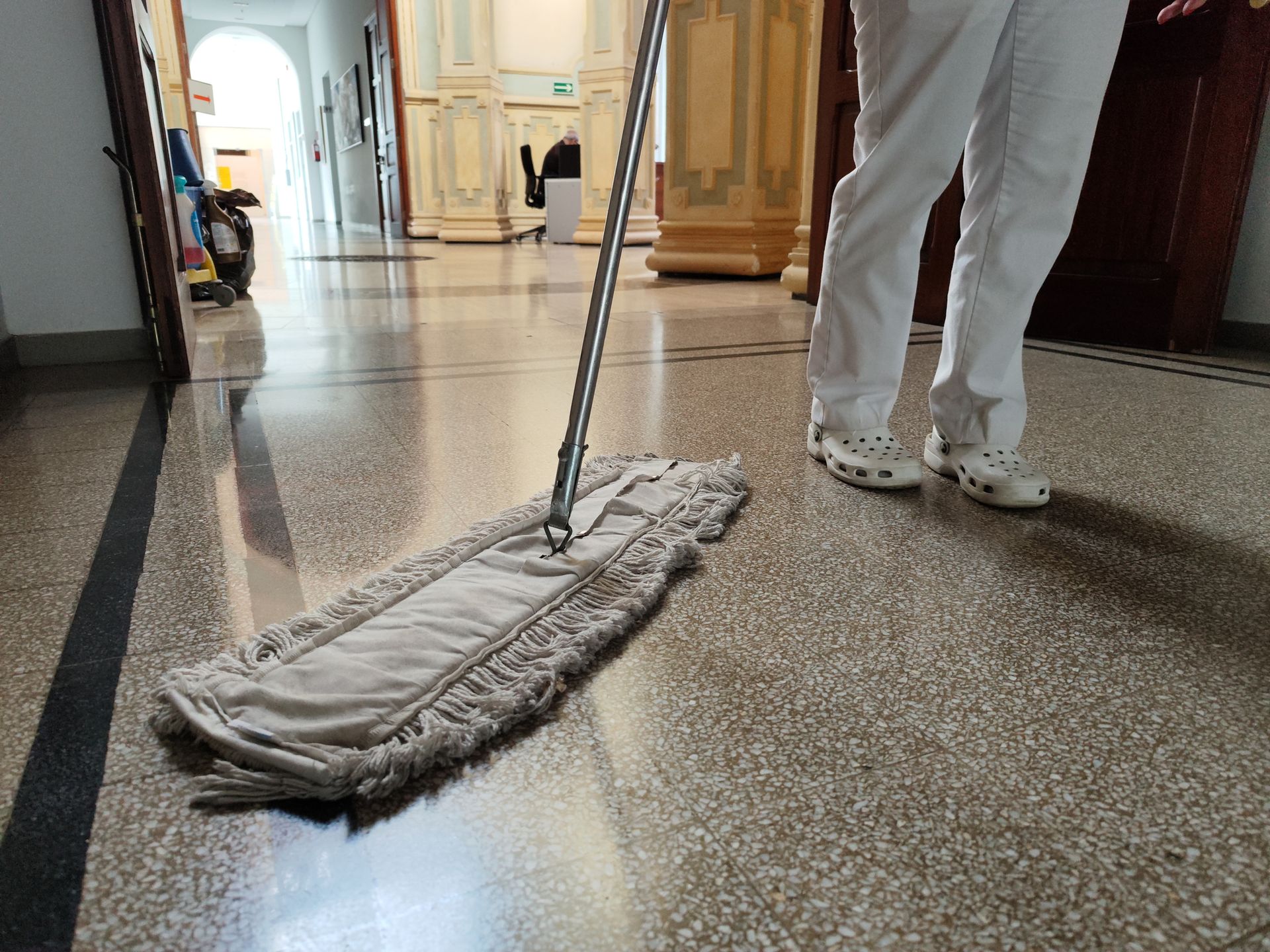 Person in white pants and clogs mopping a stone hallway with a long-handled mop.