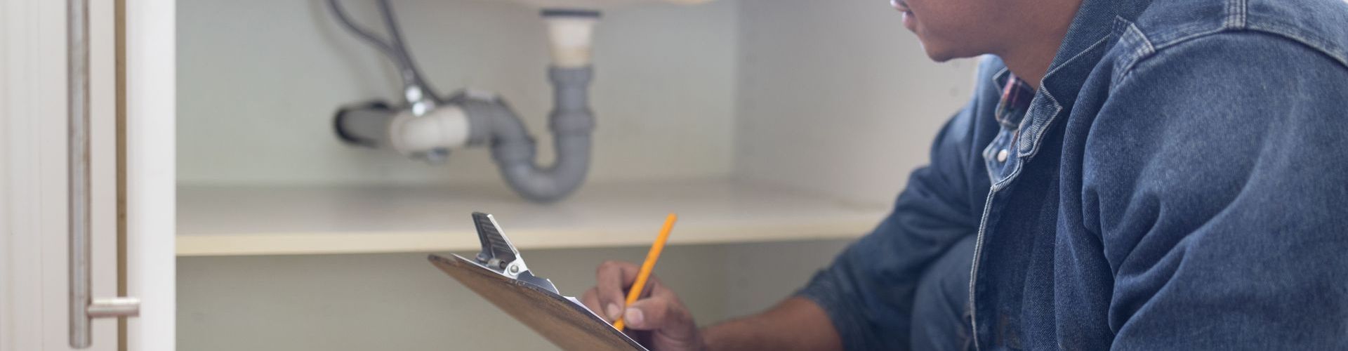 A plumber is writing on a clipboard under a sink.