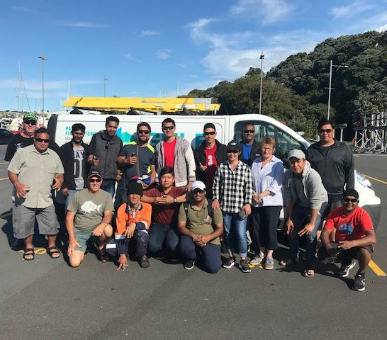 Group of people posing by a white van with a yellow ladder rack on a sunny day at a marina.