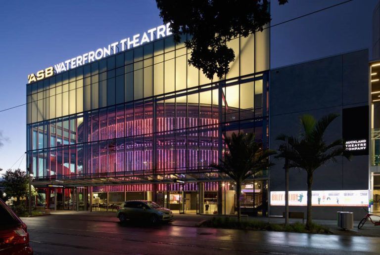 SB Waterfront Theatre illuminated at night. Large glass facade with glowing lights. A car and trees in front.