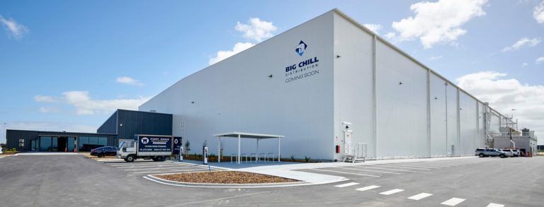 Large white industrial building with a logo on the side and a truck in the foreground, under a blue sky.