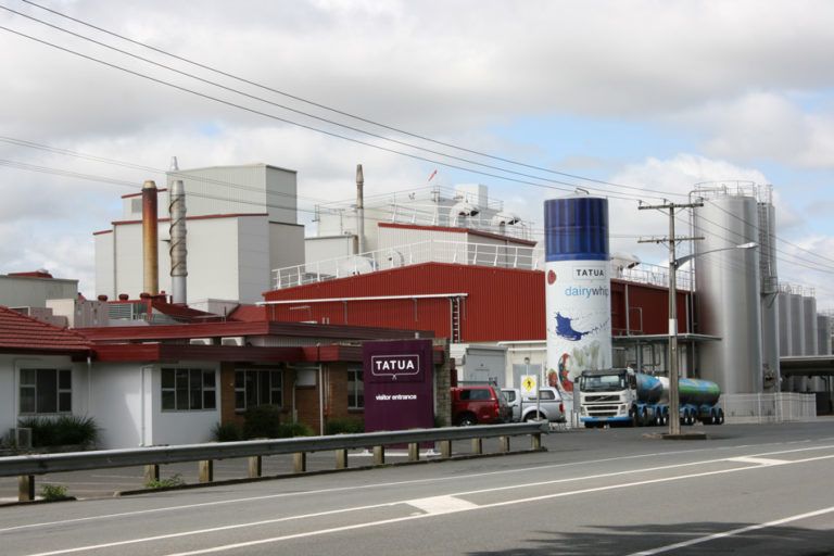 Factory complex with silos, trucks, and a sign in front of a road under a cloudy sky.