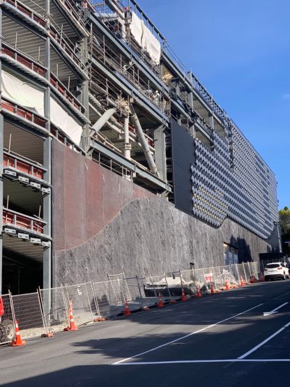 Construction site: steel-framed building with dark exterior on a sloped street, safety cones line the road.