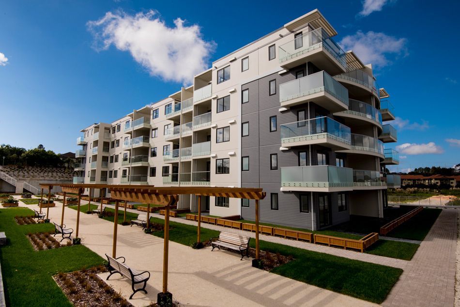 Apartment building with balconies and a pathway lined with greenery and seating, under a blue sky.