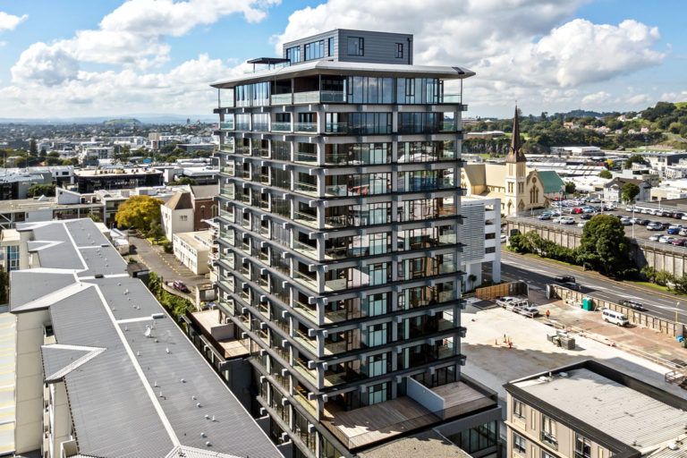 High-rise apartment building with balconies, gray frame, and glass windows, in a city with a church in the background.