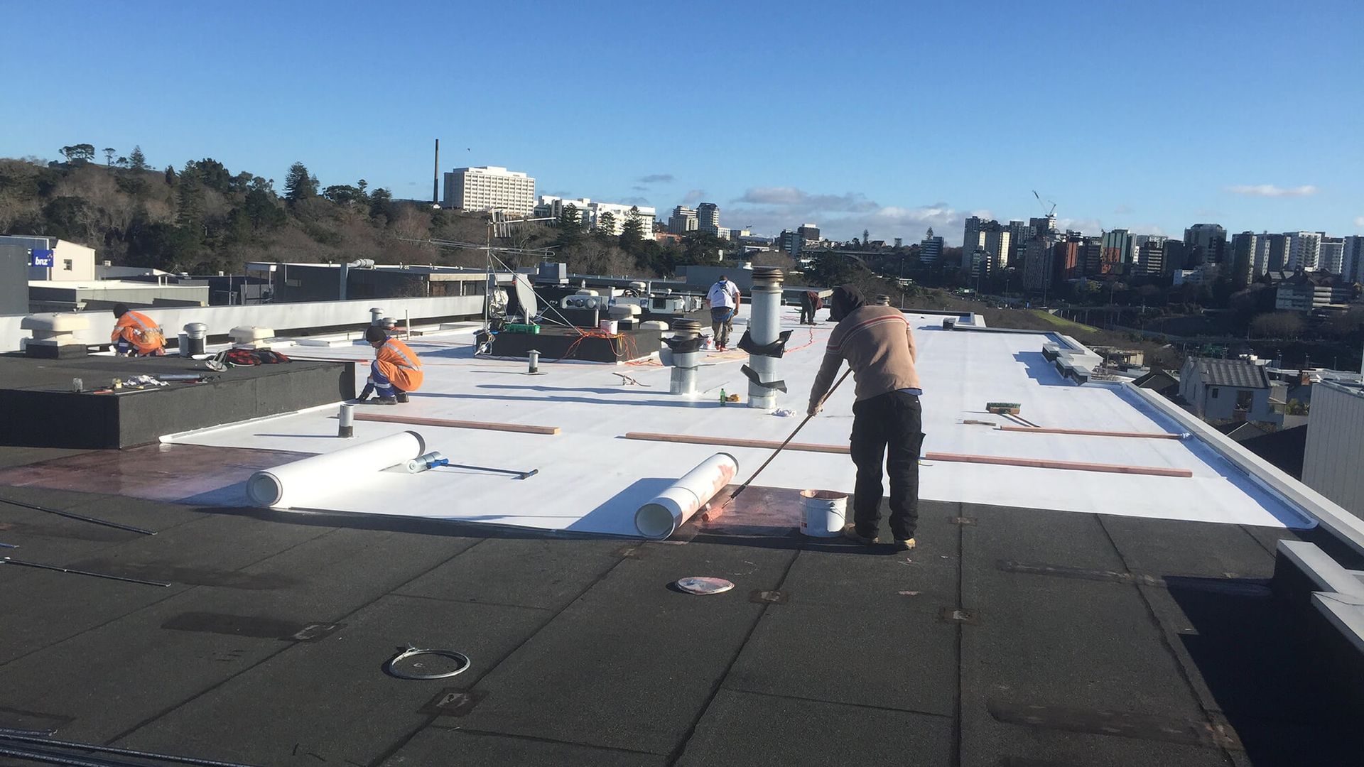 Workers installing white roofing material on a flat roof. Clear sky, cityscape in the background.