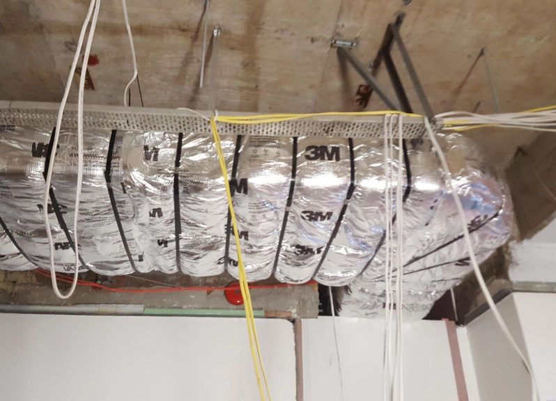 Flexible ductwork wrapped in foil insulation, suspended from a concrete ceiling with wires hanging down.