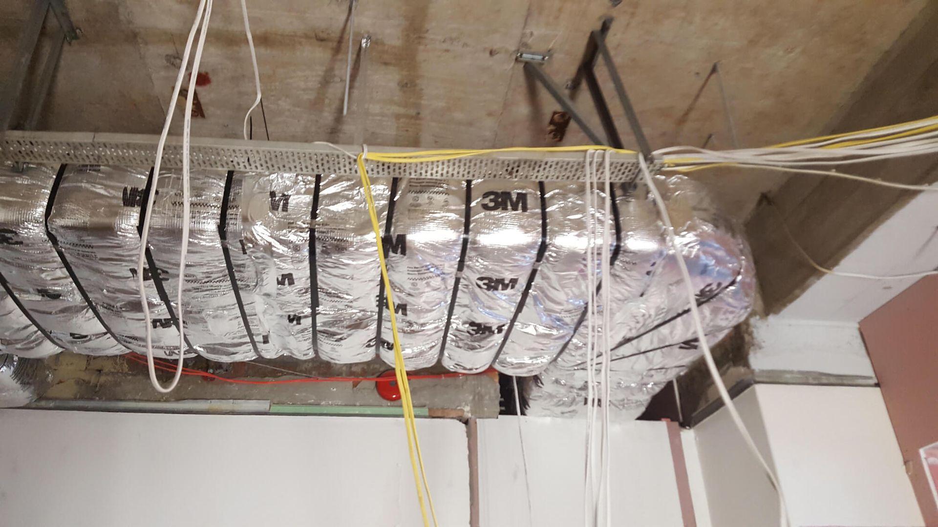 Silver ductwork wrapped in insulation and supported by wires and metal, under a ceiling.