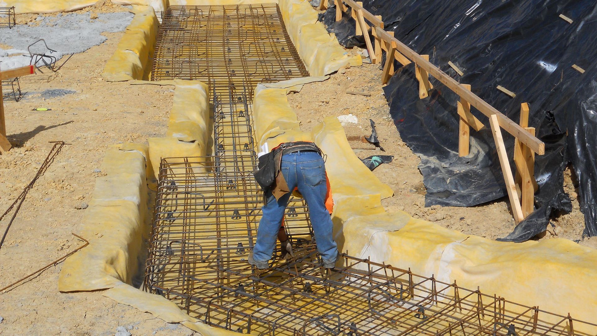 Construction worker assembling rebar framework on a hillside. Yellow fabric and wooden barriers surround the site.