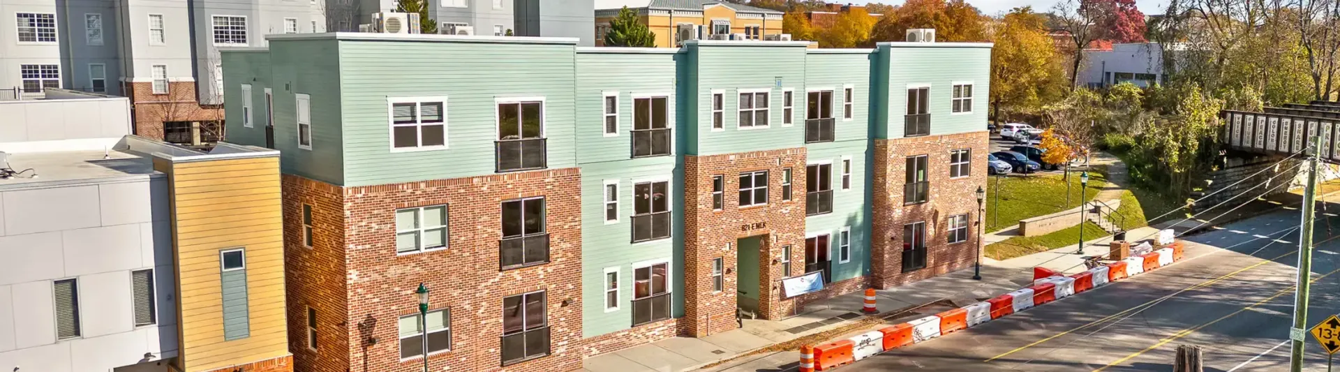 Multi-story apartment buildings with light green and stone-colored facades. A street is in front, with construction barriers.