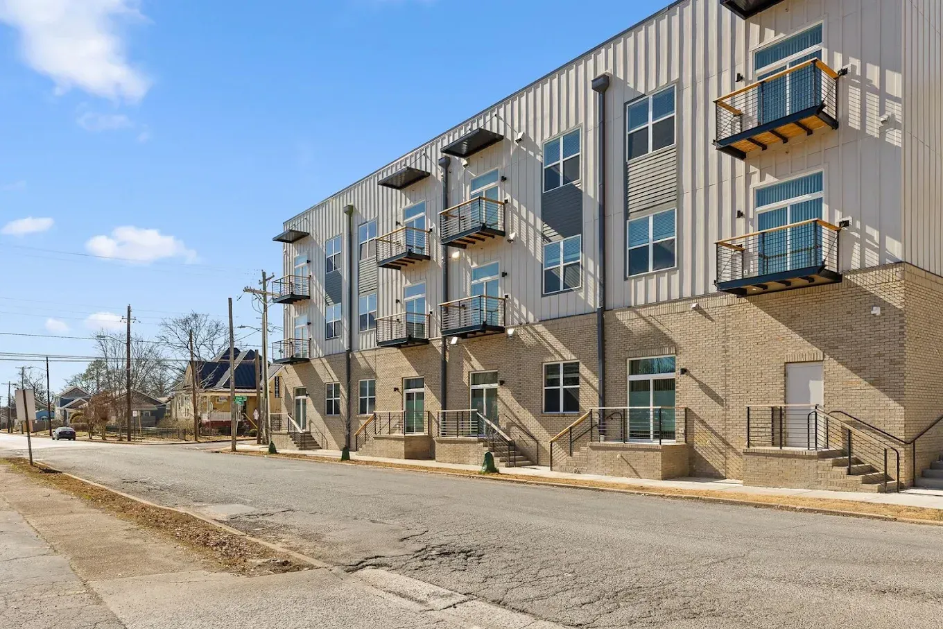 Multi-story building with metal siding and small balconies. Brick base, street in foreground, sunny day.