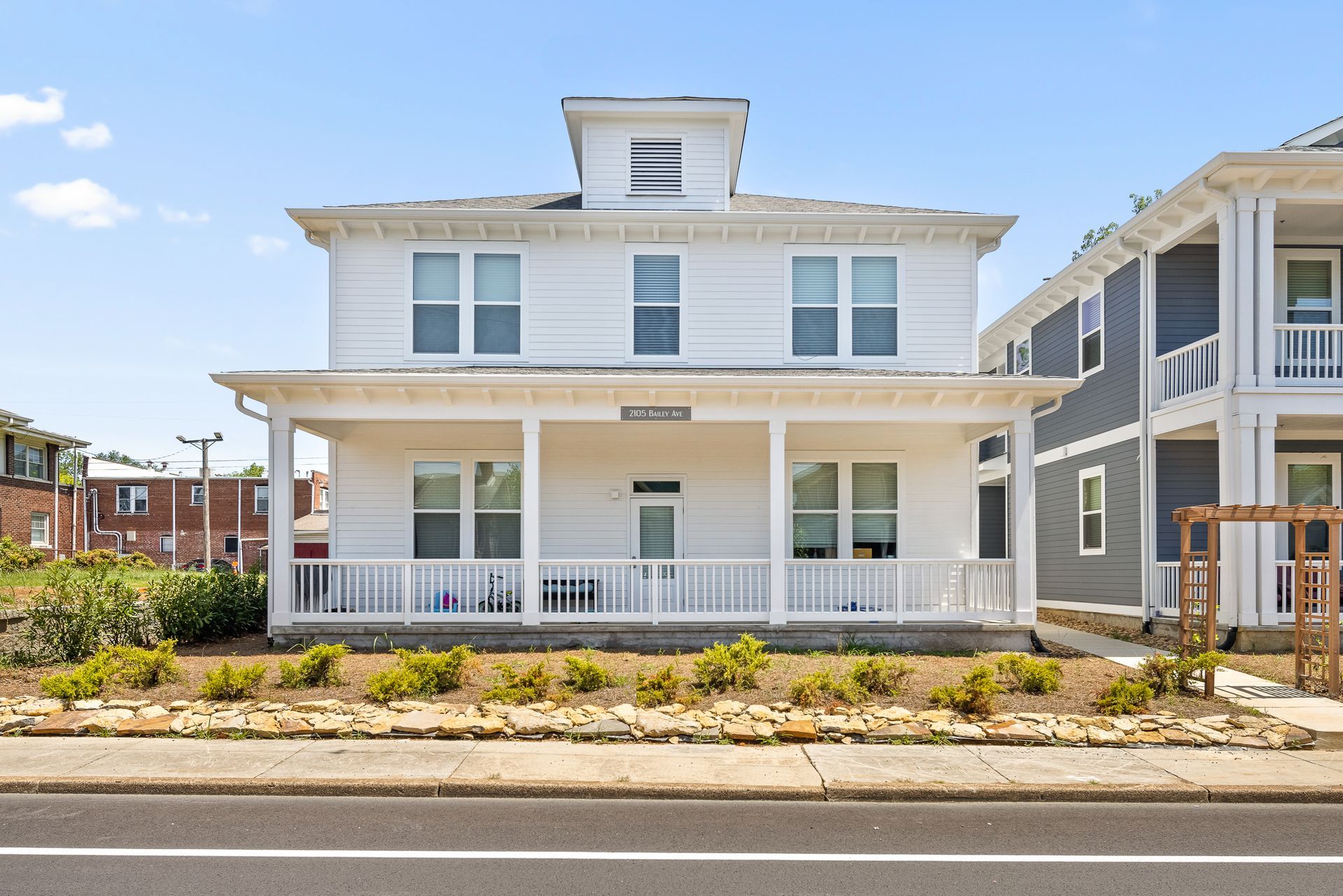 White two-story rental complex with a porch and dormer. It is in front of a road with other buildings nearby.