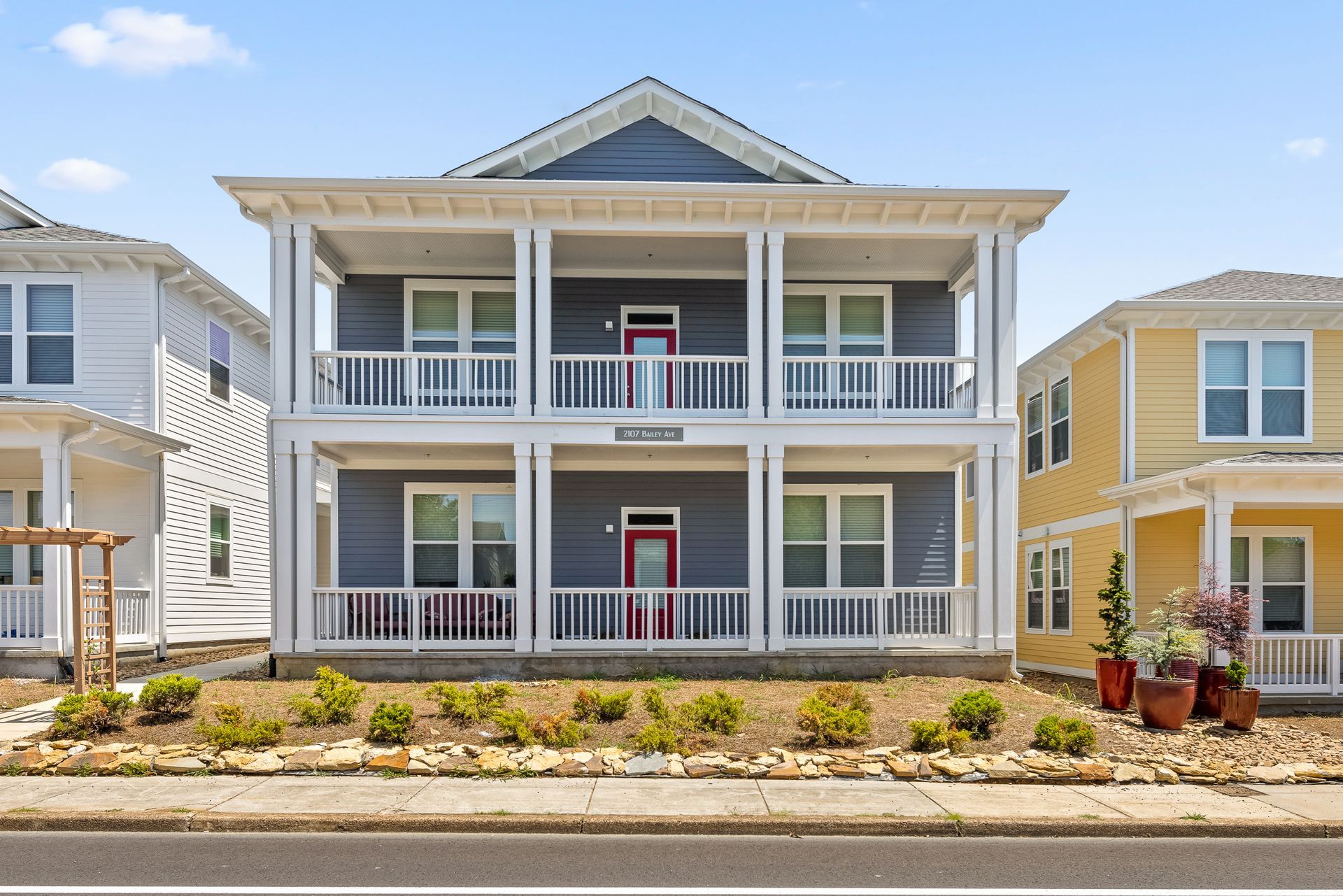 Two-story blue house with white columns and railings. Red front doors, and a low rock bed in front of the building.