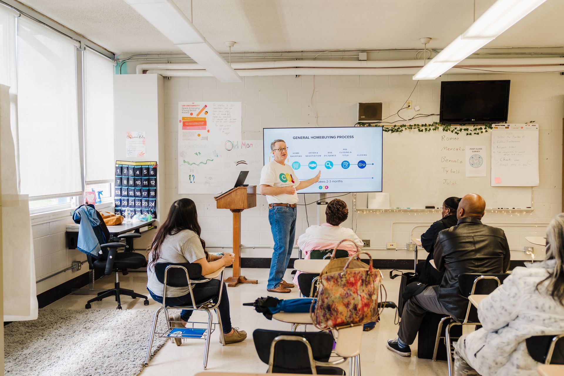 A man gives a presentation in a classroom with several seated attendees. He stands near a screen showing education materials.