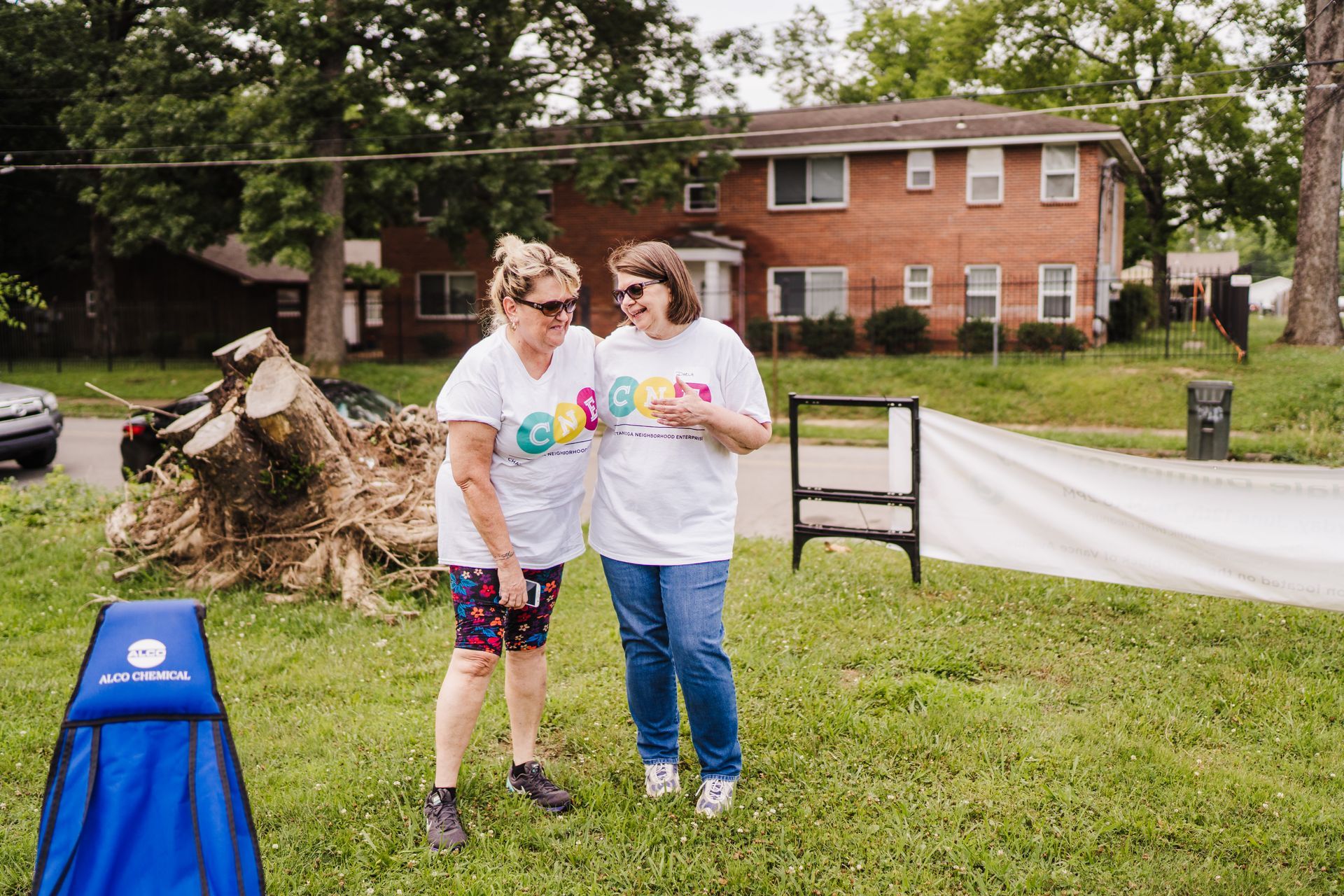 Two women wearing white shirts with cne logos stand on grass, talking. A brick building and tree debris are in the background.