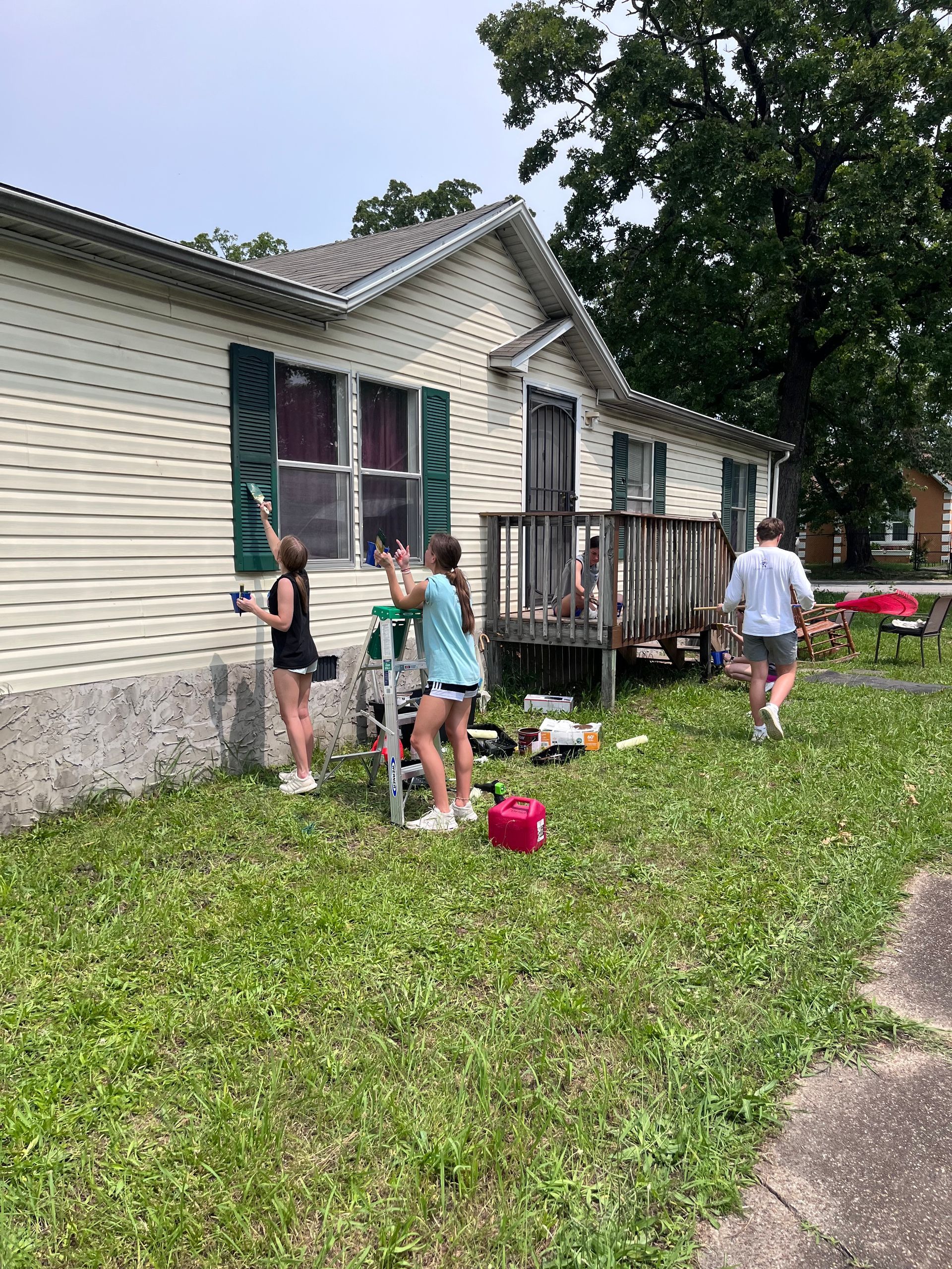 People paint exterior shutters on a house. Grass and supplies are in the yard.