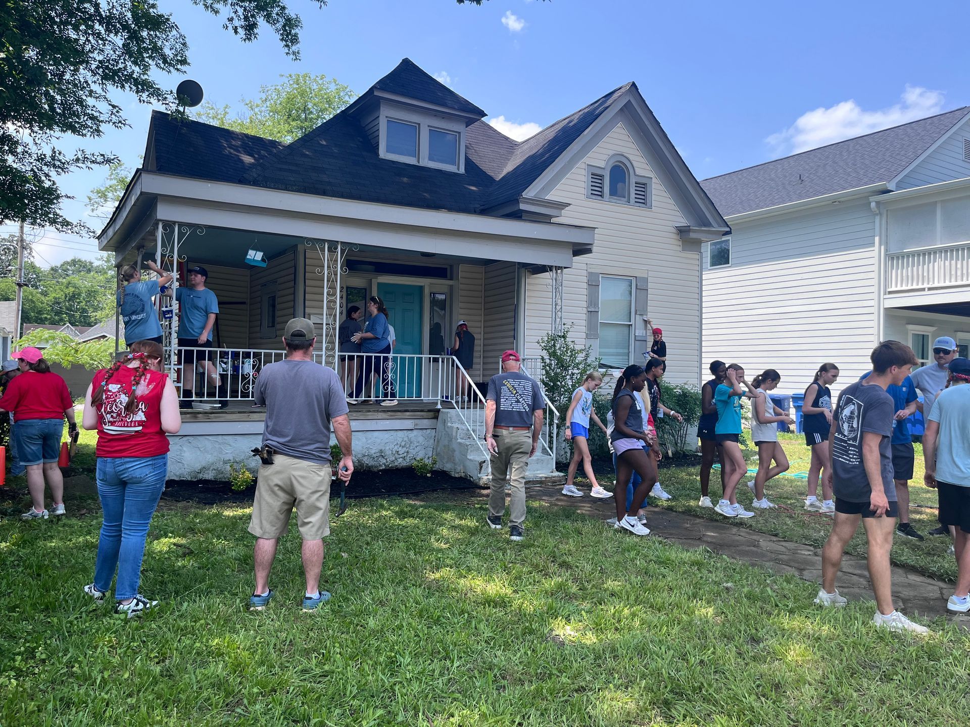 People gathered in front of a house, possibly filming. A crew on the porch, others on the lawn, bright day.