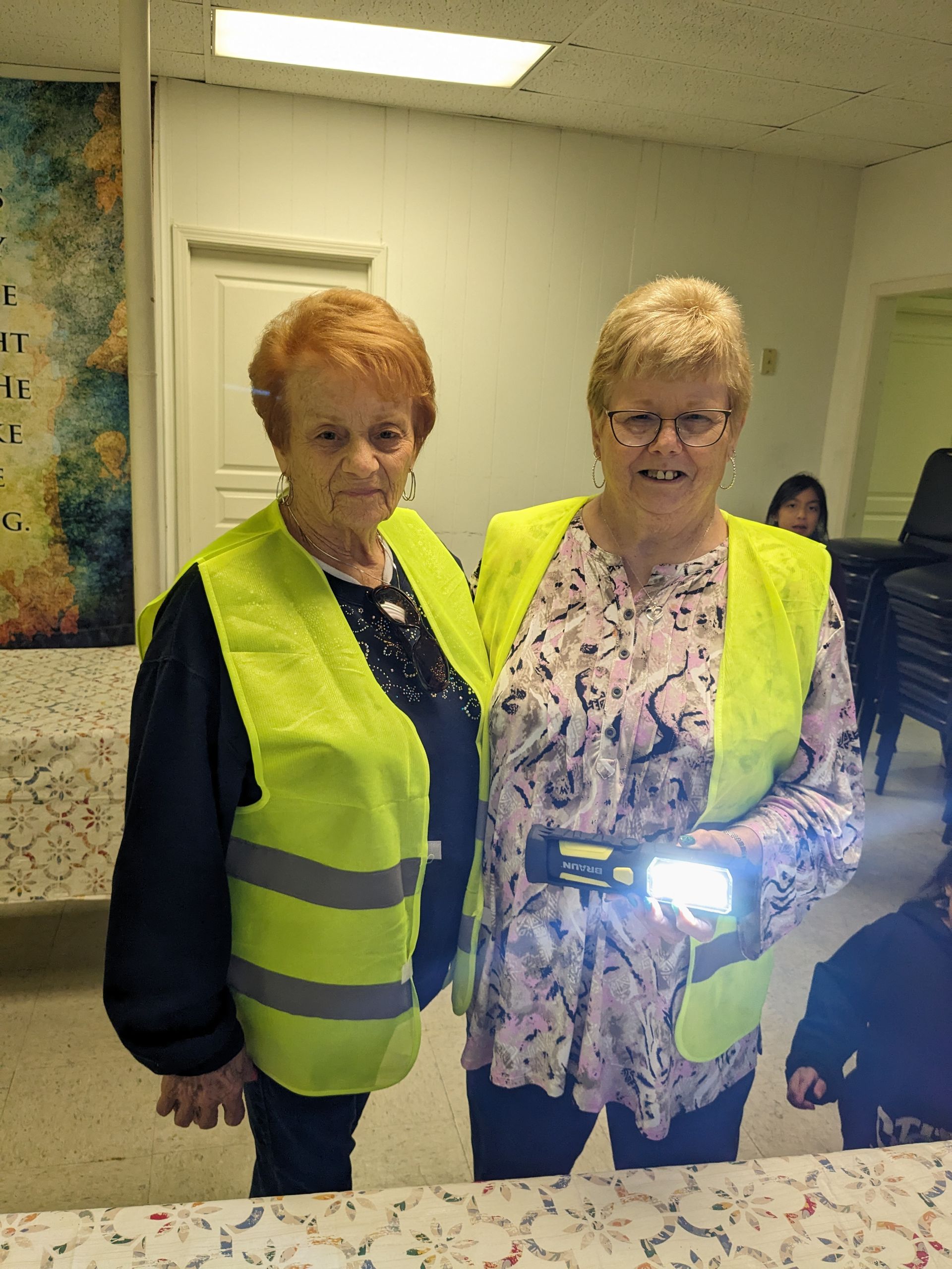 Two women wearing safety vests indoors, one holding a flashlight.