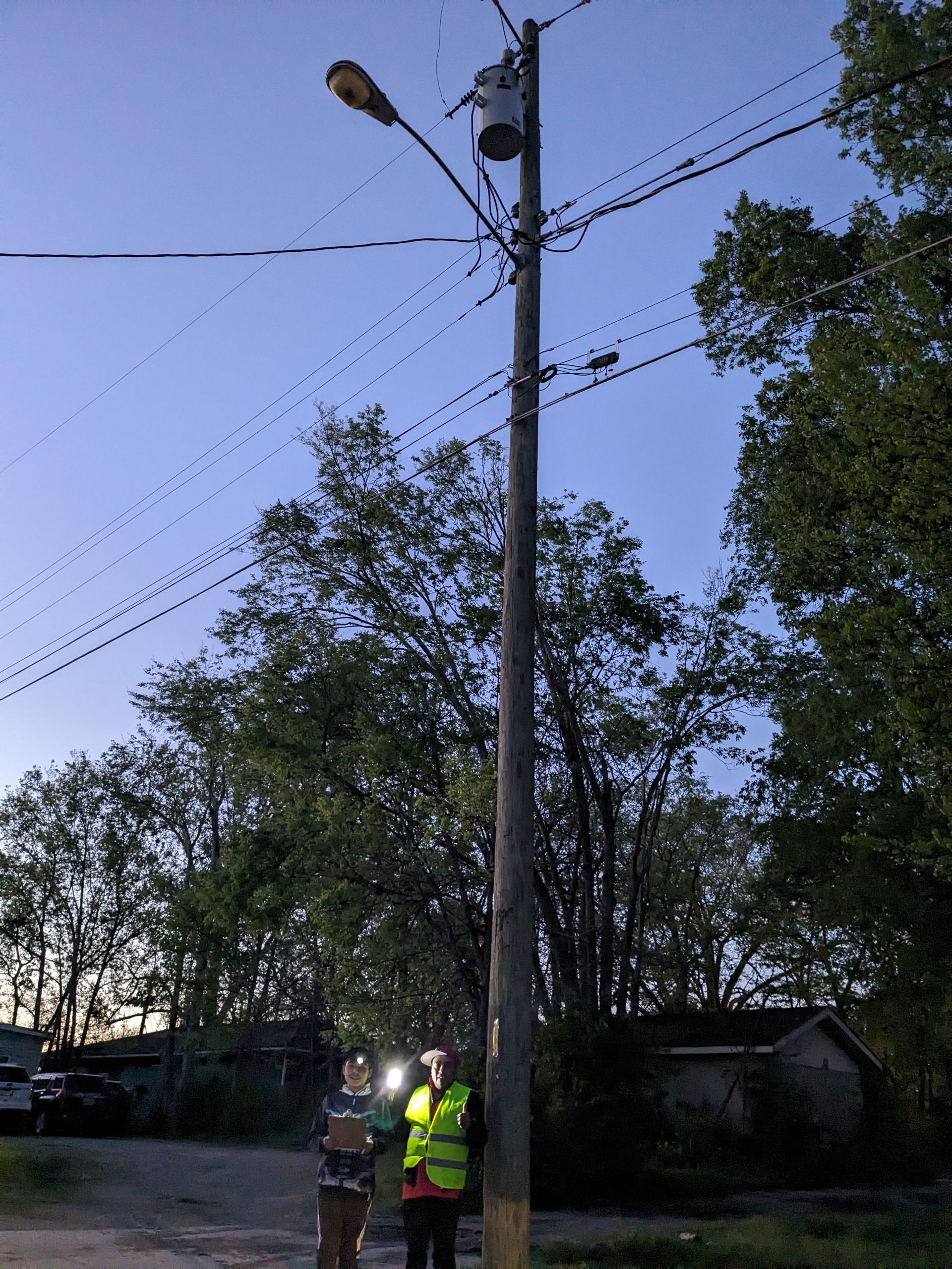 Two people in safety vests stand near a utility pole with a street light. Dusk setting.