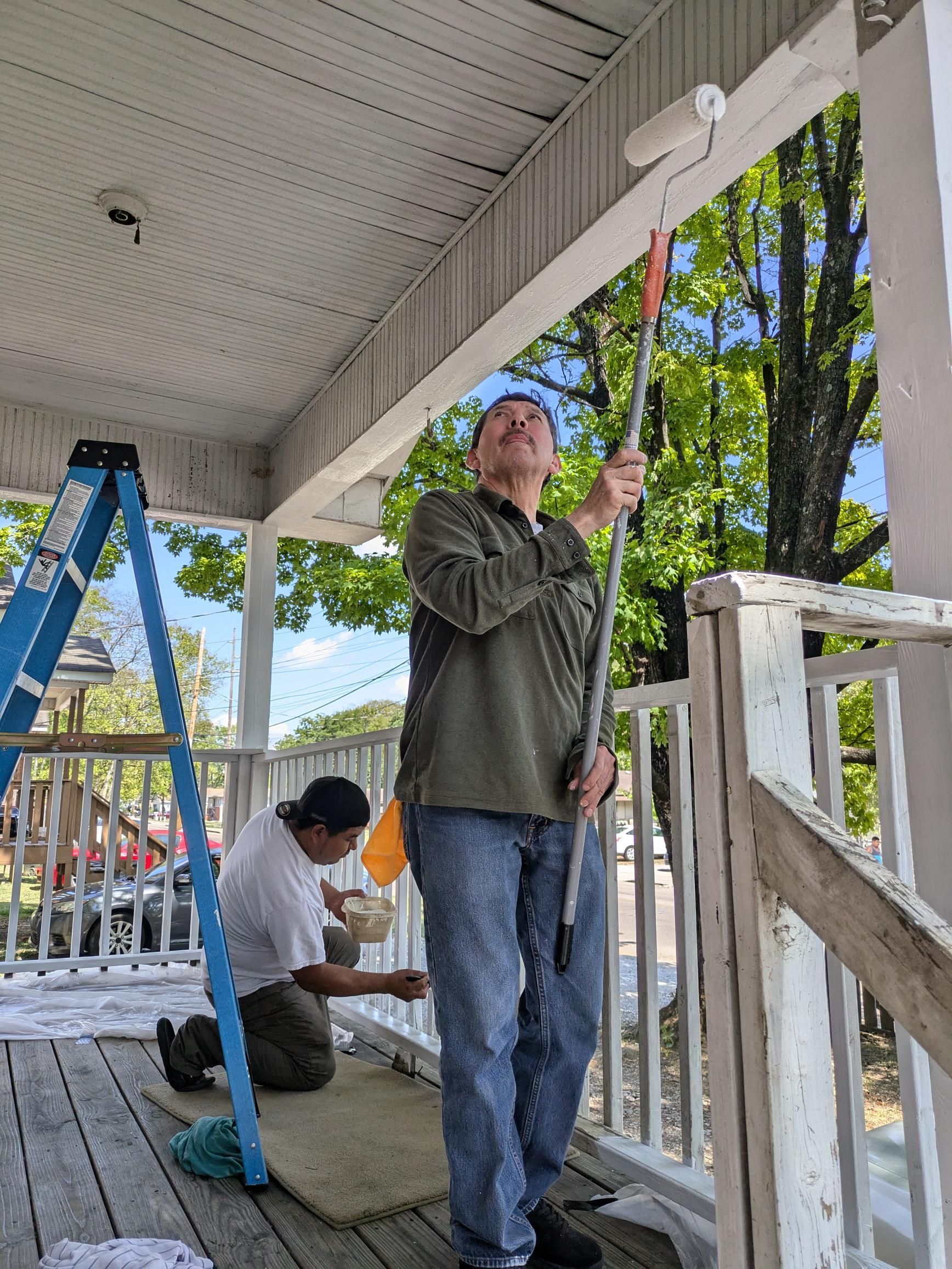 Two people painting a white porch. One on a ladder, the other using a roller, outdoors, trees in background.